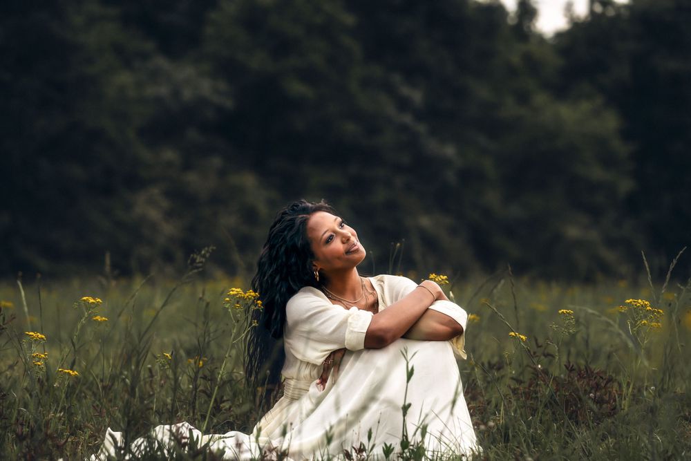 Woman in white dress sitting peacefully in tall grass field surrounded by yellow wildflowers.