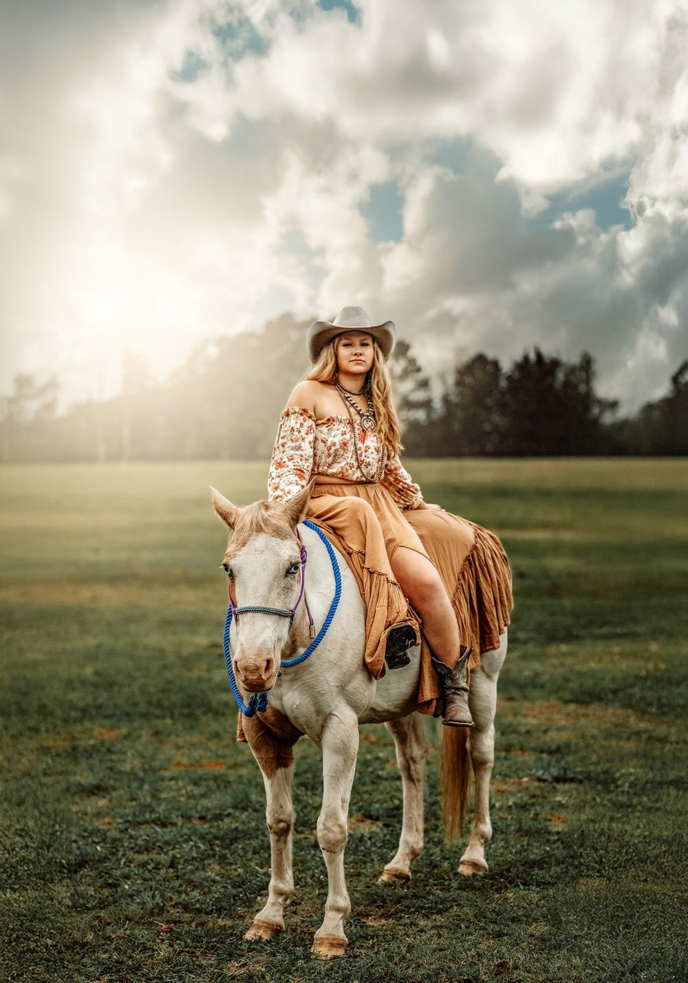 Young woman in cowboy hat and boho outfit riding a white horse in a green pasture.