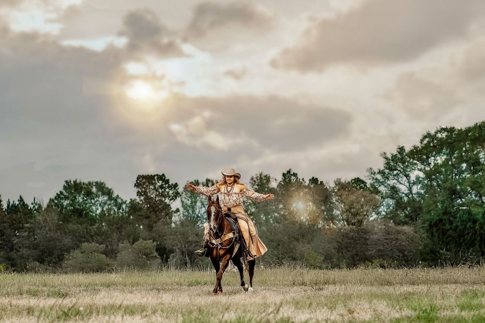 Cowgirl riding a dark horse across an open field under a dramatic cloudy sunset sky.