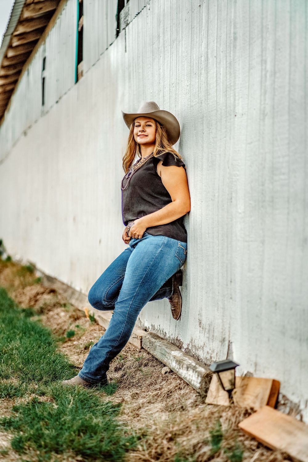 Young woman in cowboy hat, jeans, and boots leaning against a barn wall.