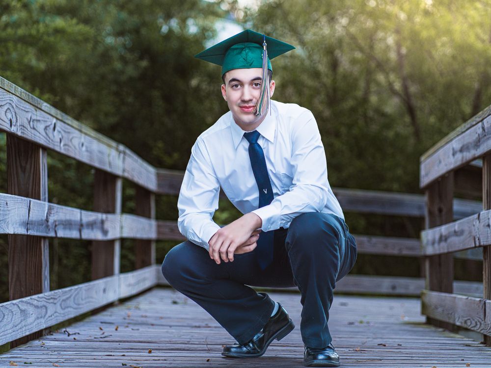 Young male graduate in green cap crouching on a wooden bridge surrounded by trees.