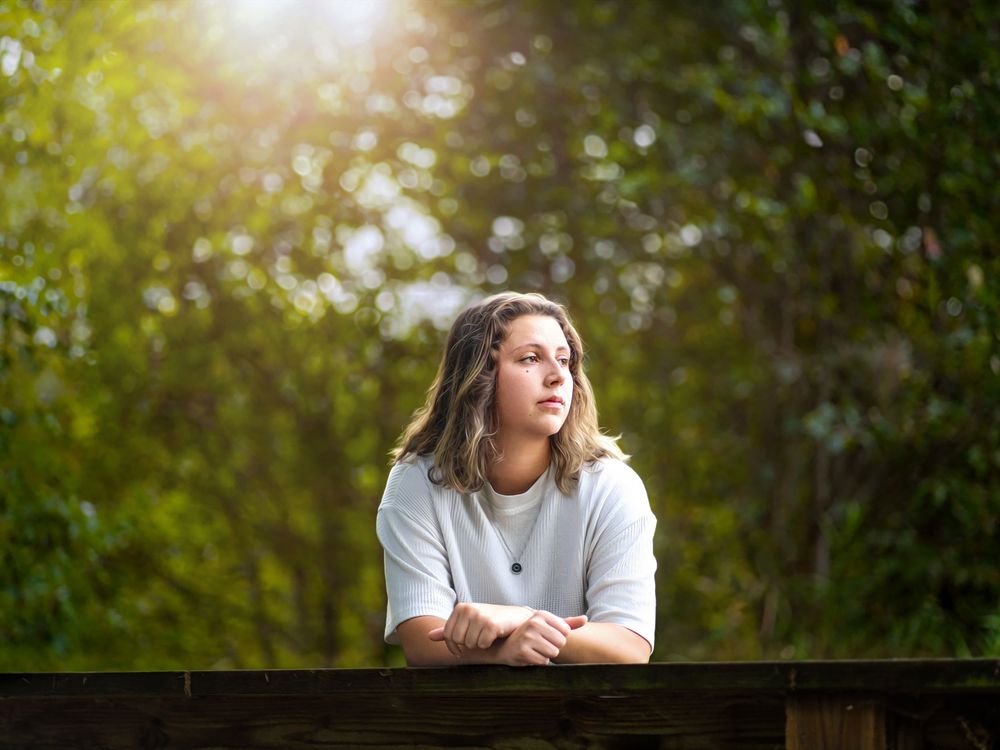 Young woman in white sweater leaning on a wooden railing surrounded by sunlit trees.
