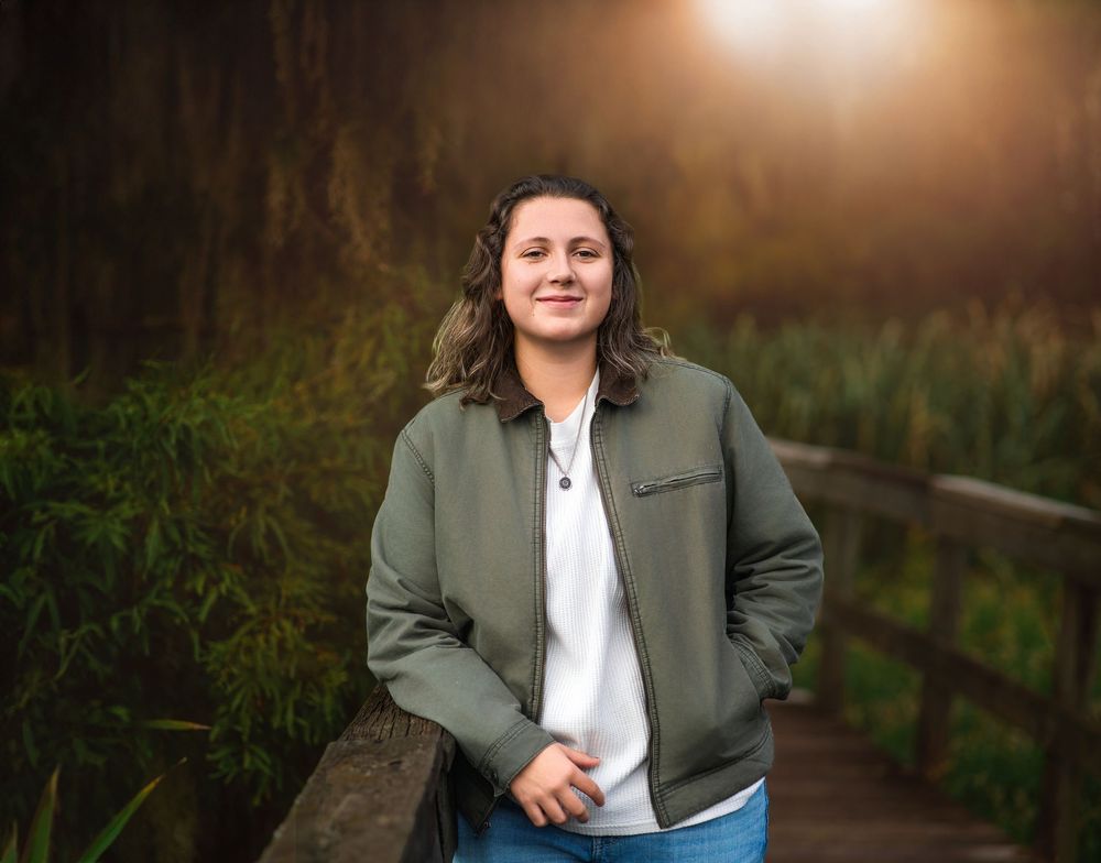 Smiling young woman in green bomber jacket standing on a boardwalk at golden hour.