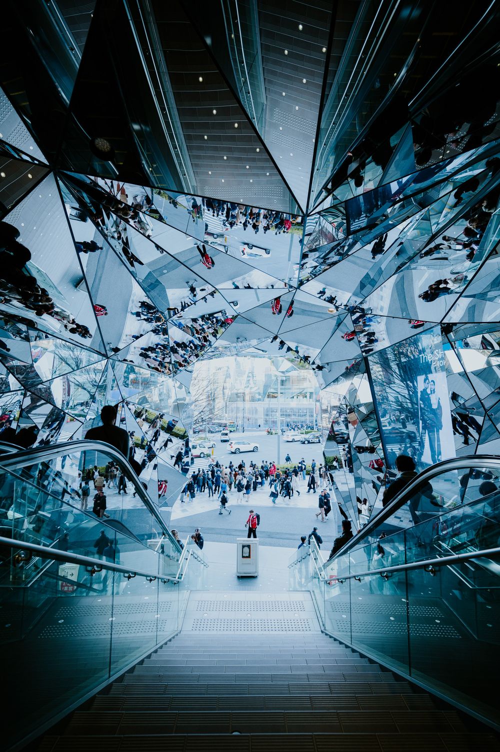 Futuristic mirrored interior of a modern shopping mall or transit station with reflective ceiling panels creating kaleidoscopic patterns.