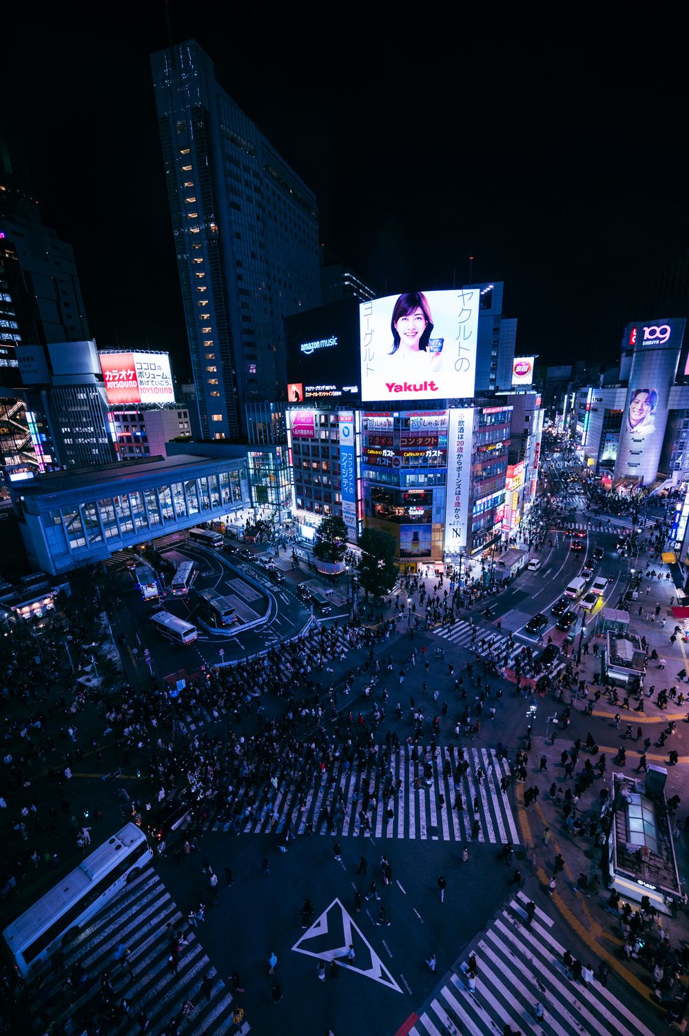 Aerial view of the famous Shibuya crossing in Tokyo at night with crowds of people and illuminated billboards.