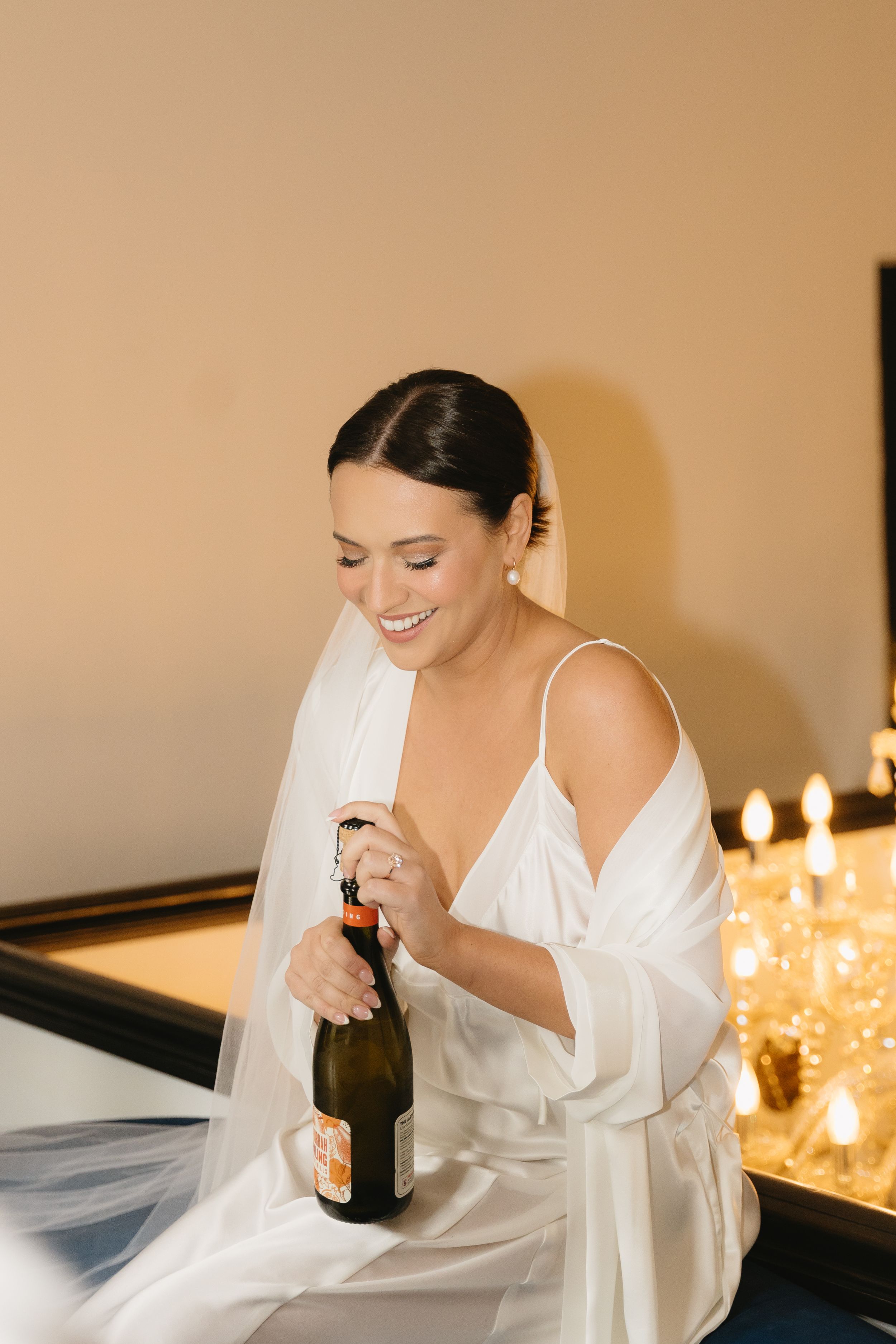 A person in a white robe sits elegantly while holding a bottle in a warmly lit room.