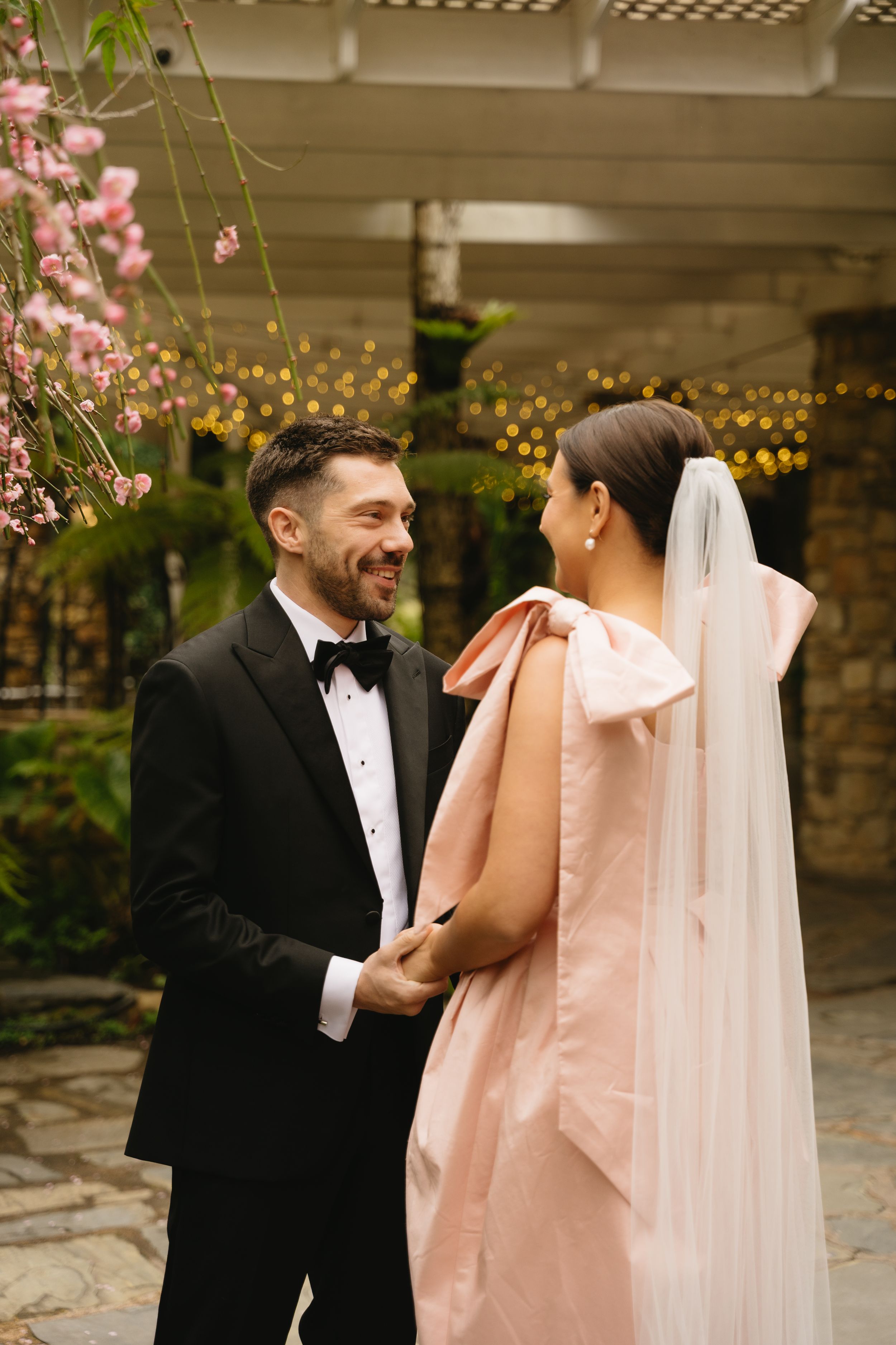 A couple shares a romantic moment outdoors surrounded by pink cherry blossoms and string lights.