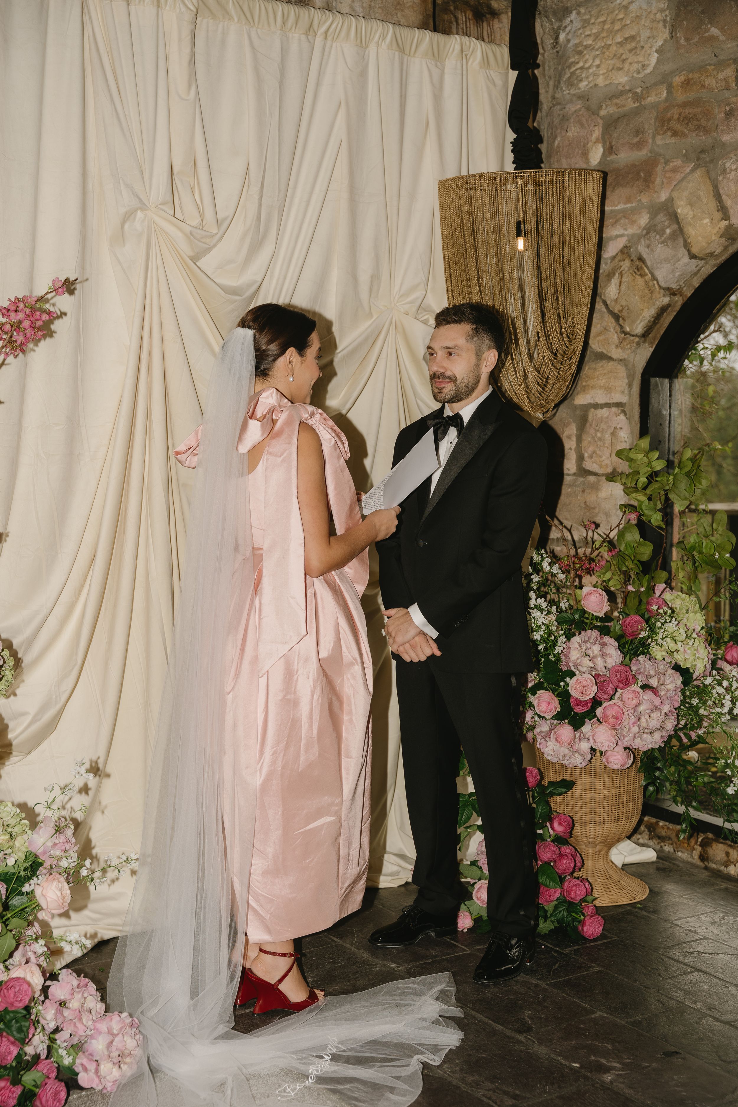 A romantic wedding scene with pink florals and white draping against a stone wall backdrop.