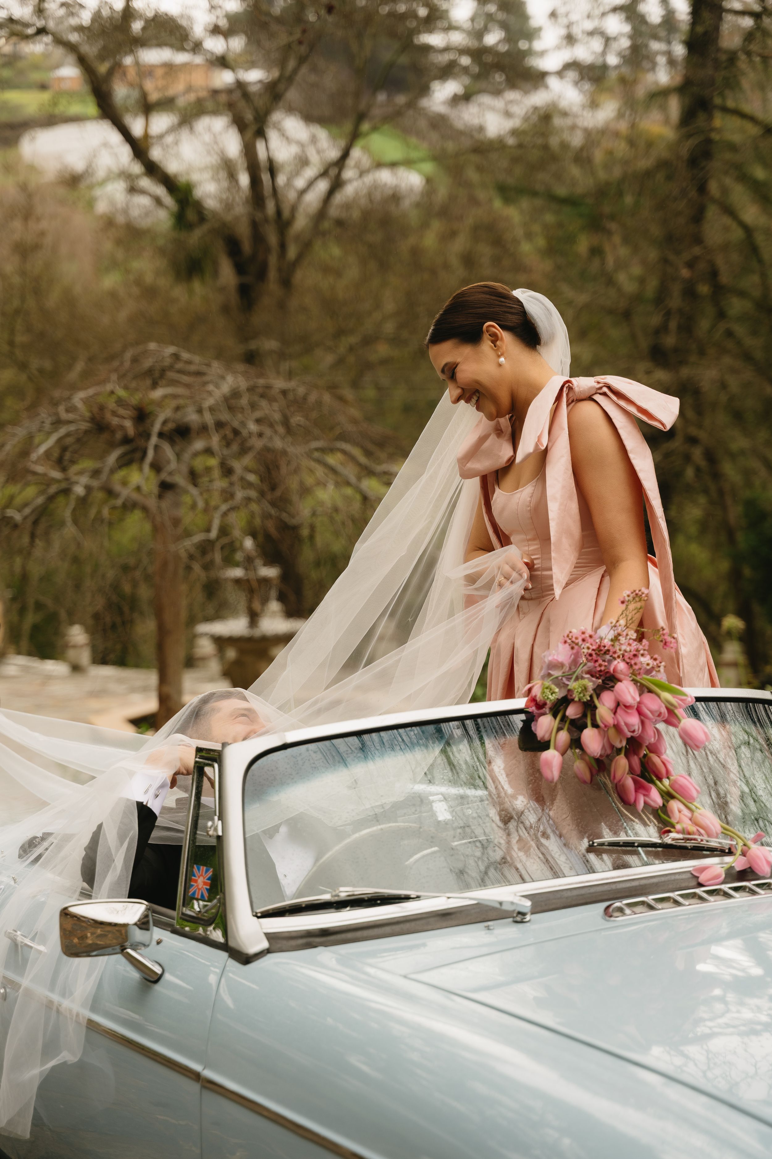 Elegant wedding scene with bride in pink dress leaning over vintage convertible car.