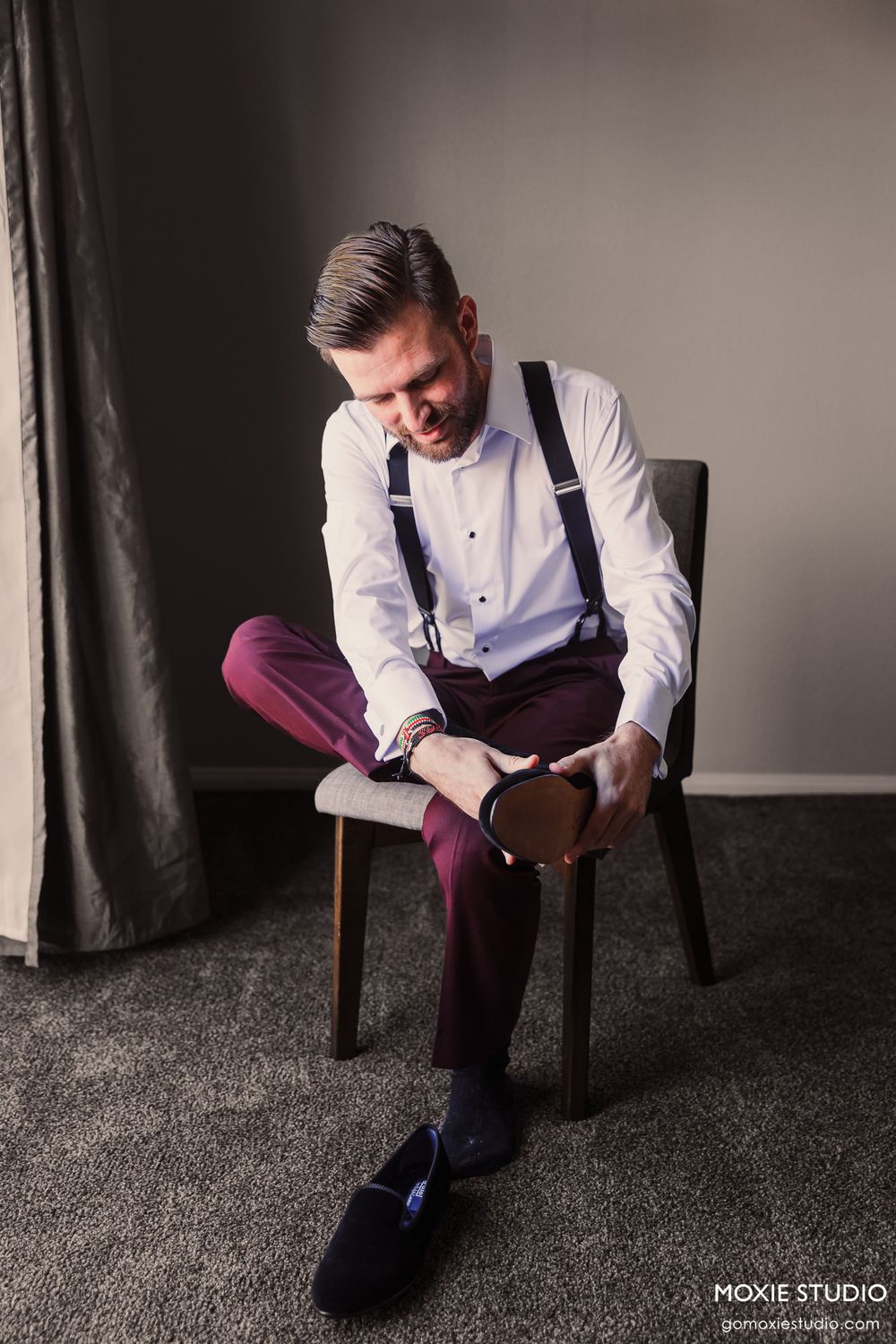 Groom in burgundy pants and white shirt sitting on chair in dramatic lighting.