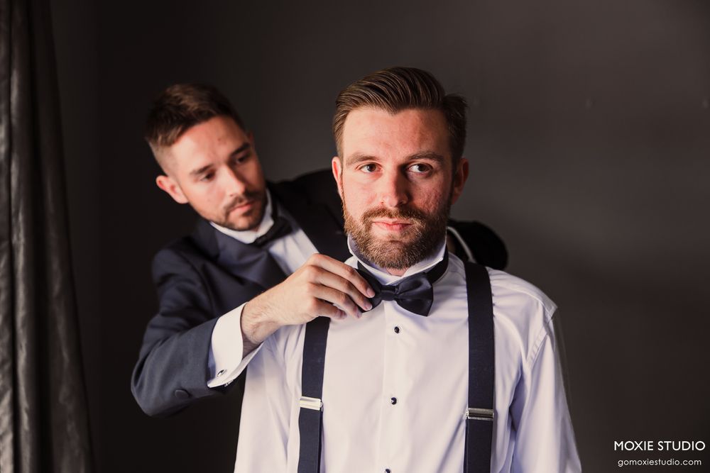 A groomsman helps adjust the bow tie of a groom wearing suspenders and a white dress shirt.