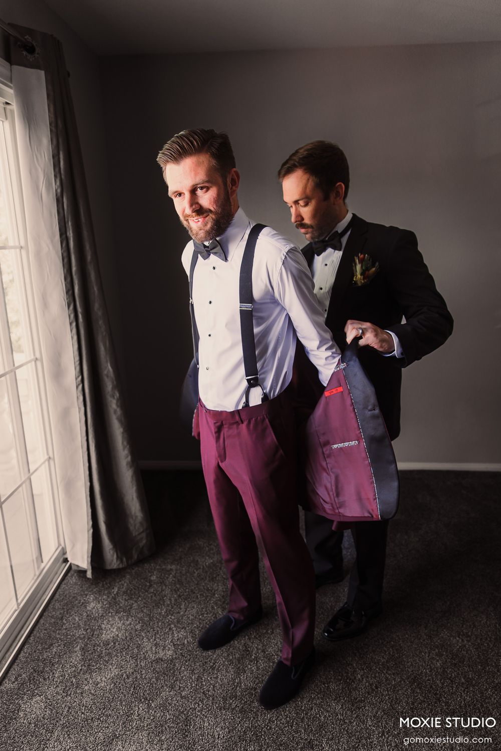 Two men in formal attire getting ready, one wearing burgundy pants and suspenders.