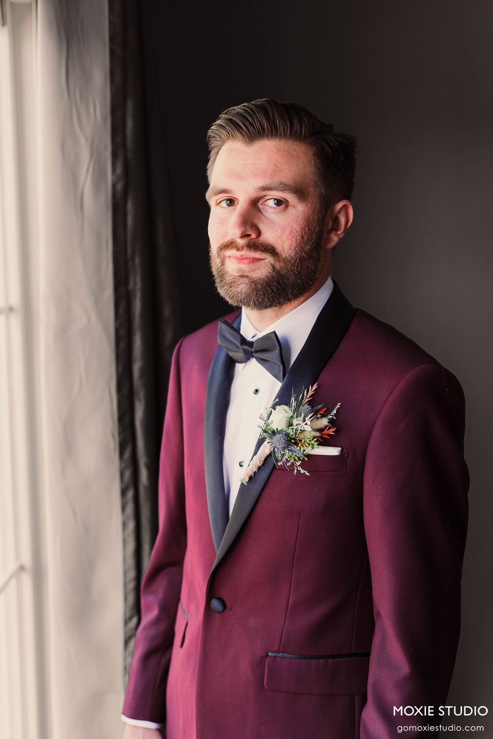 A formal portrait showing a burgundy tuxedo jacket with boutonniere by a window.