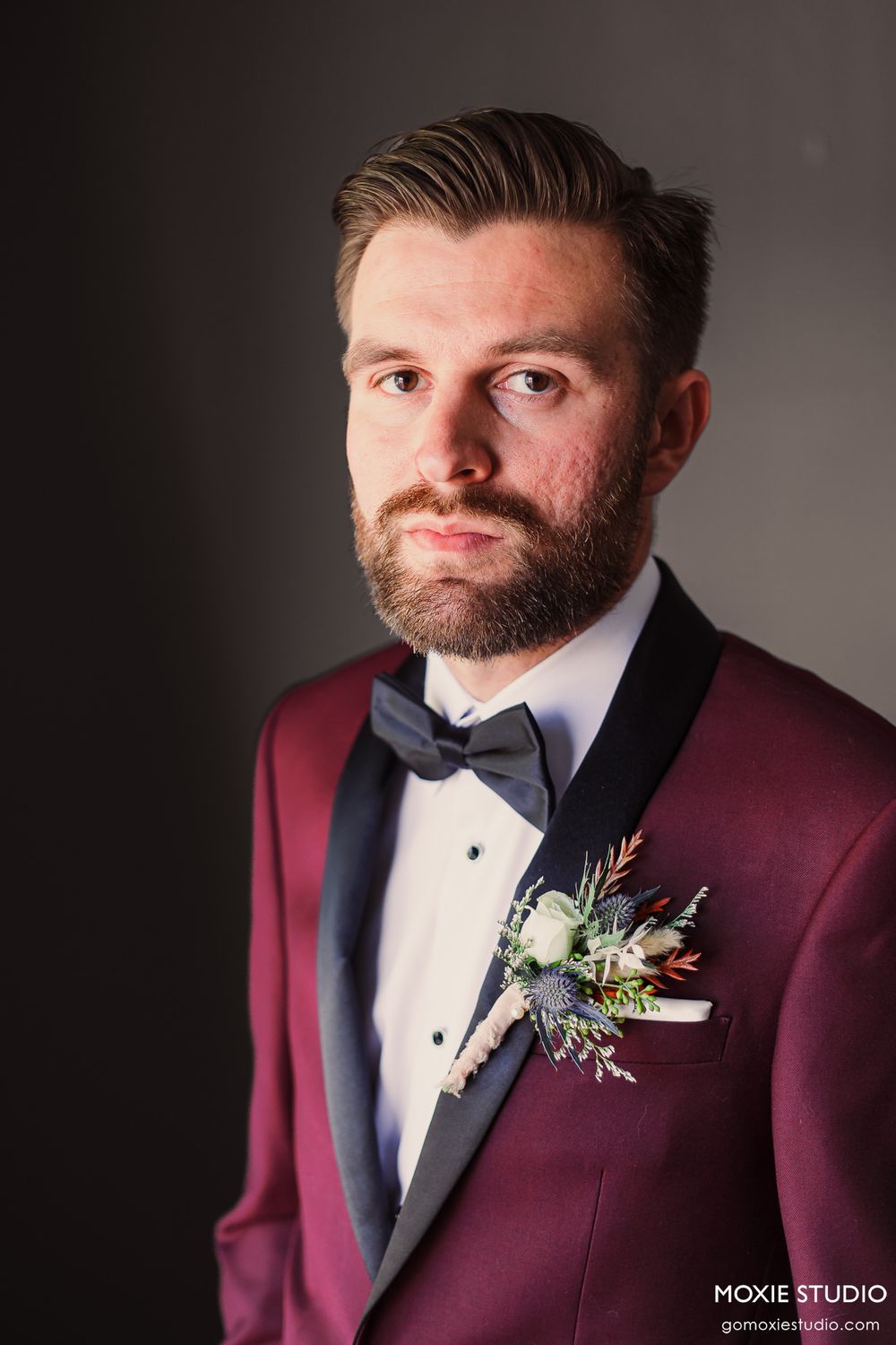 Groom in burgundy tuxedo jacket with boutonniere poses against dark background in formal portrait series.