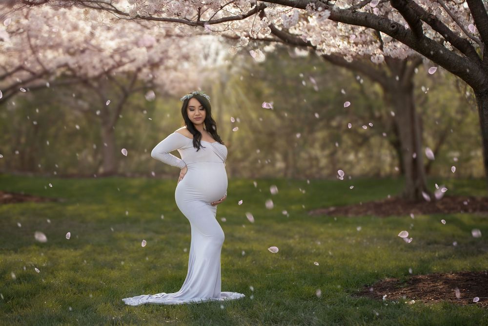 A photographer captures multiple maternity photos in white dress under cherry blossoms in a spring garden setting.