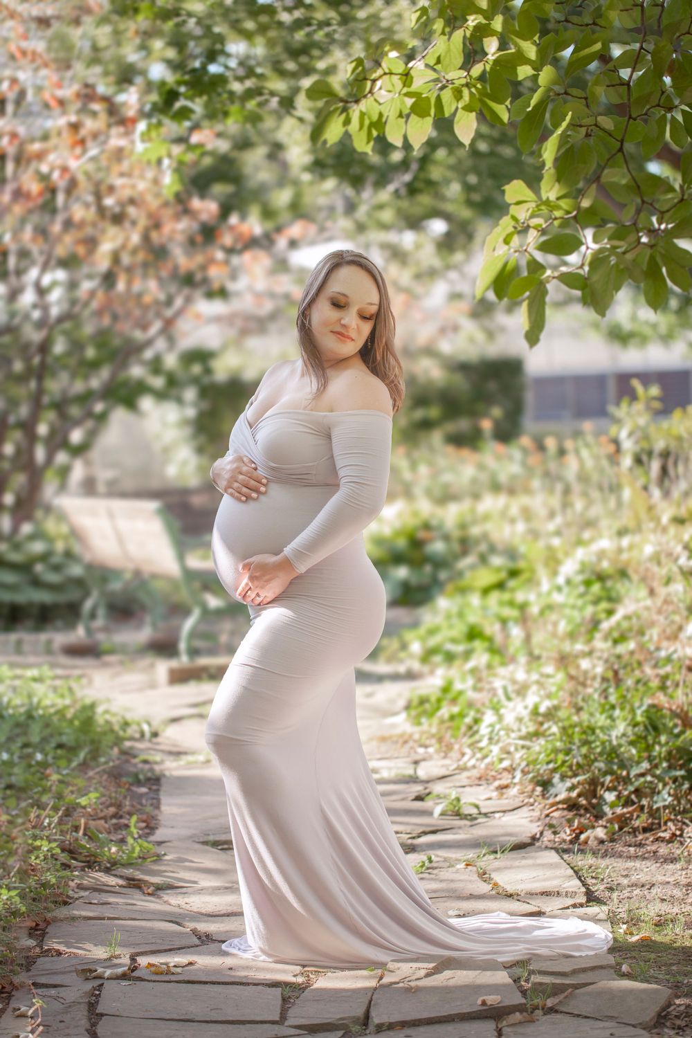 Elegant maternity photoshoot in a garden setting with soft natural lighting and stone pathway backdrop.
