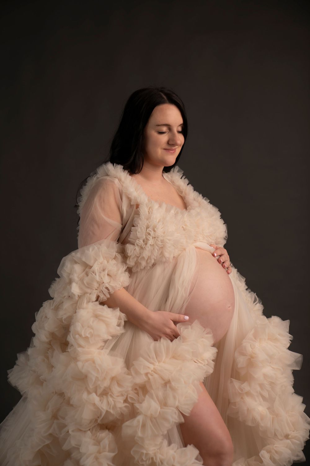 A maternity photo session featuring flowing beige tulle fabric in a dramatic ethereal style against a dark background.