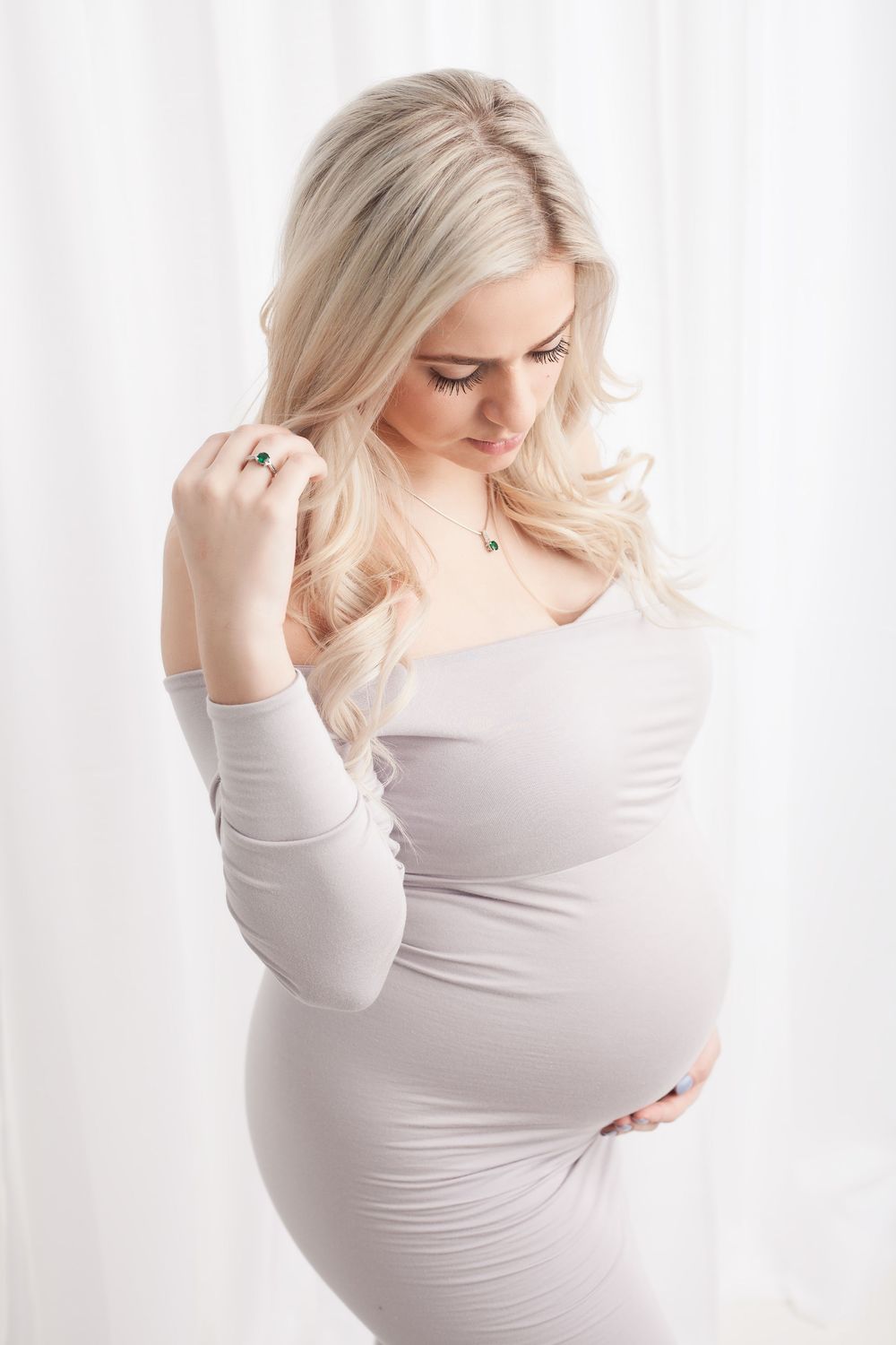 A woman in soft white clothing poses against a bright background during a gentle maternity photoshoot.