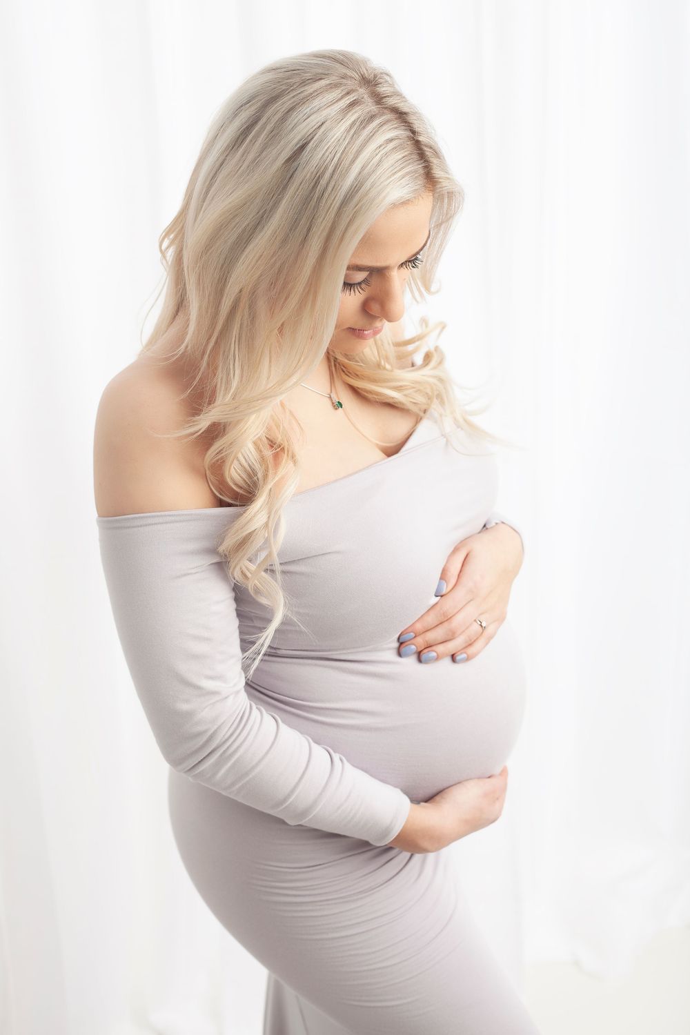 Artistic maternity photography series showing woman in light grey dress cradling baby bump against white background.