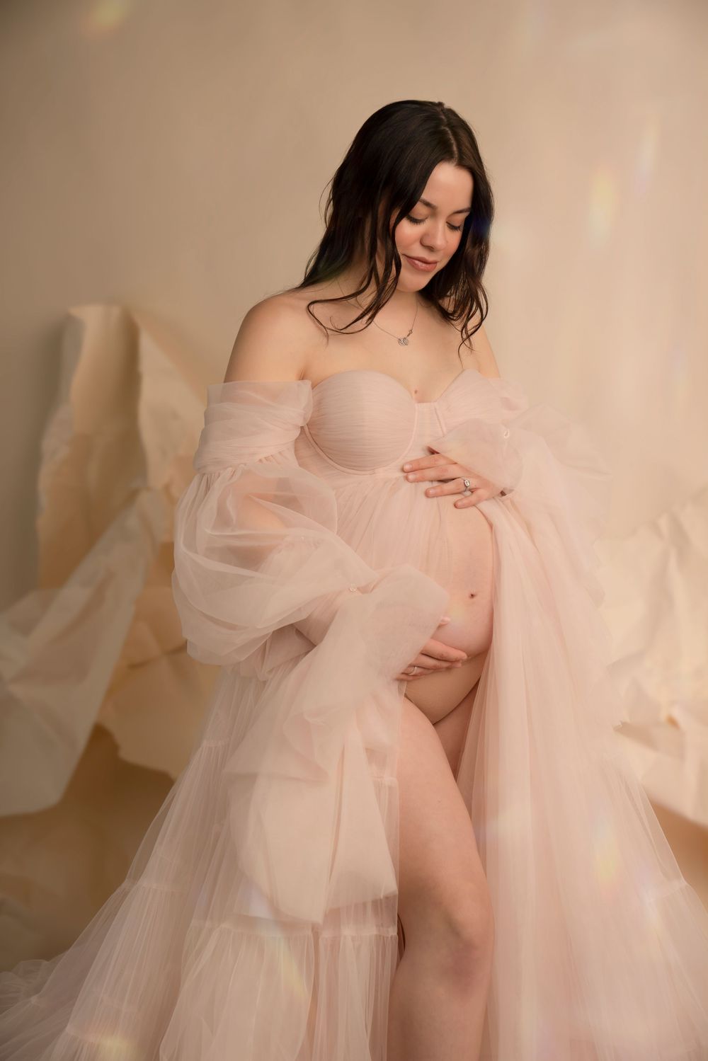 A maternity photoshoot features a person in a flowing sheer pink tulle robe against a light neutral backdrop.