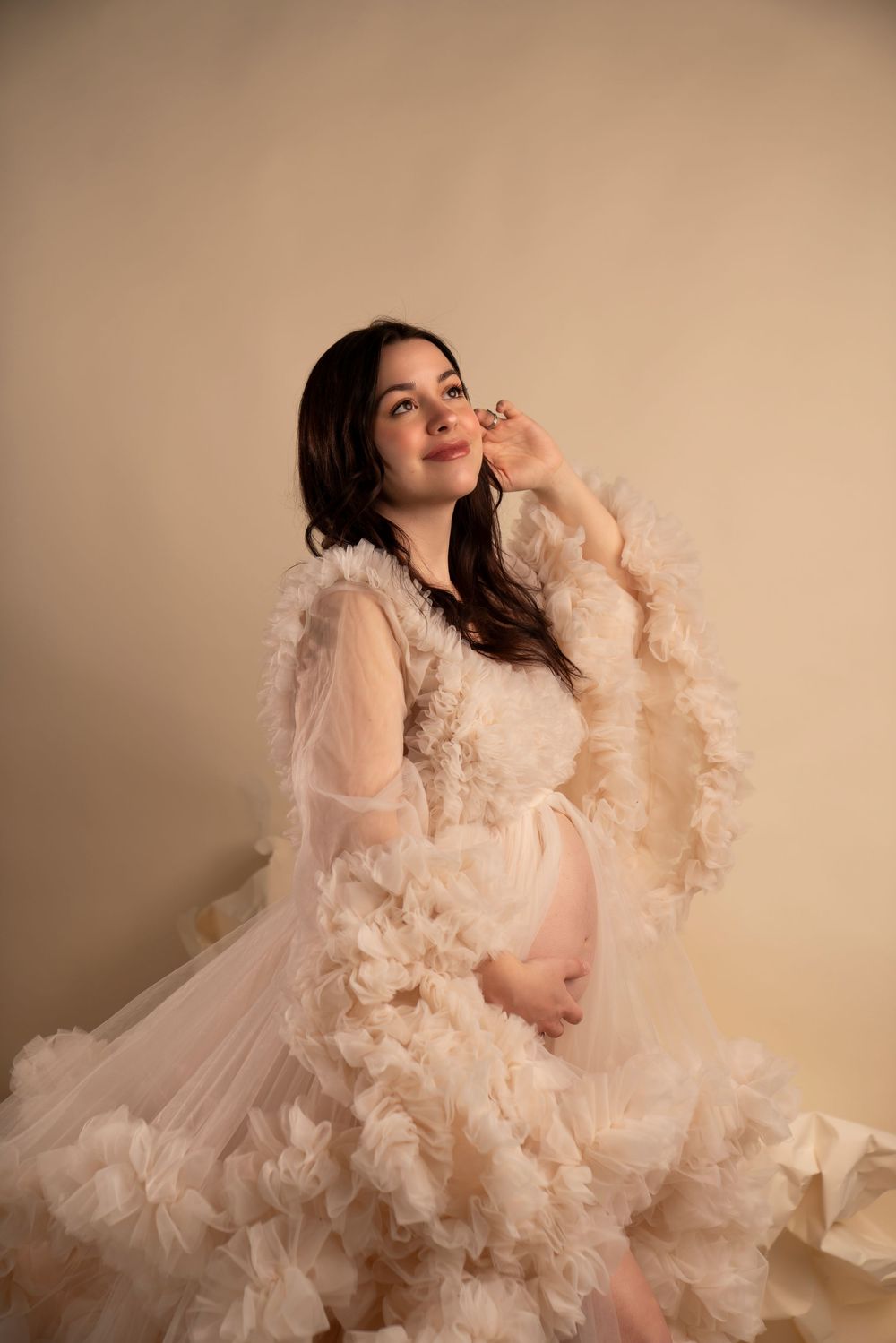 A maternity photoshoot featuring ethereal cream-colored ruffled tulle gowns against a soft neutral background.