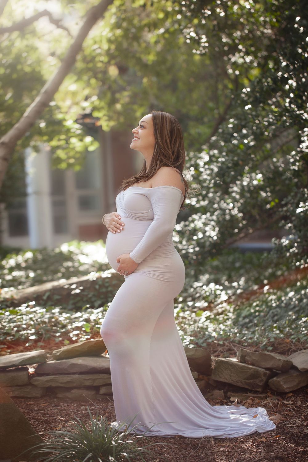 A pregnant woman in a flowing white dress poses gracefully in a sunlit garden setting with trees and rocks.