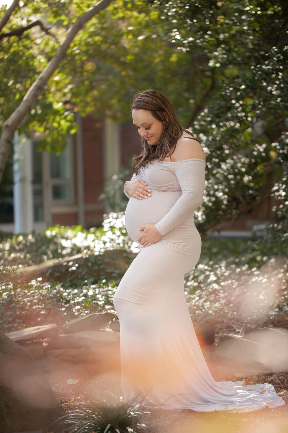 A pregnant woman in a white dress poses gracefully in a sunlit garden with delicate white flowers blooming around her.