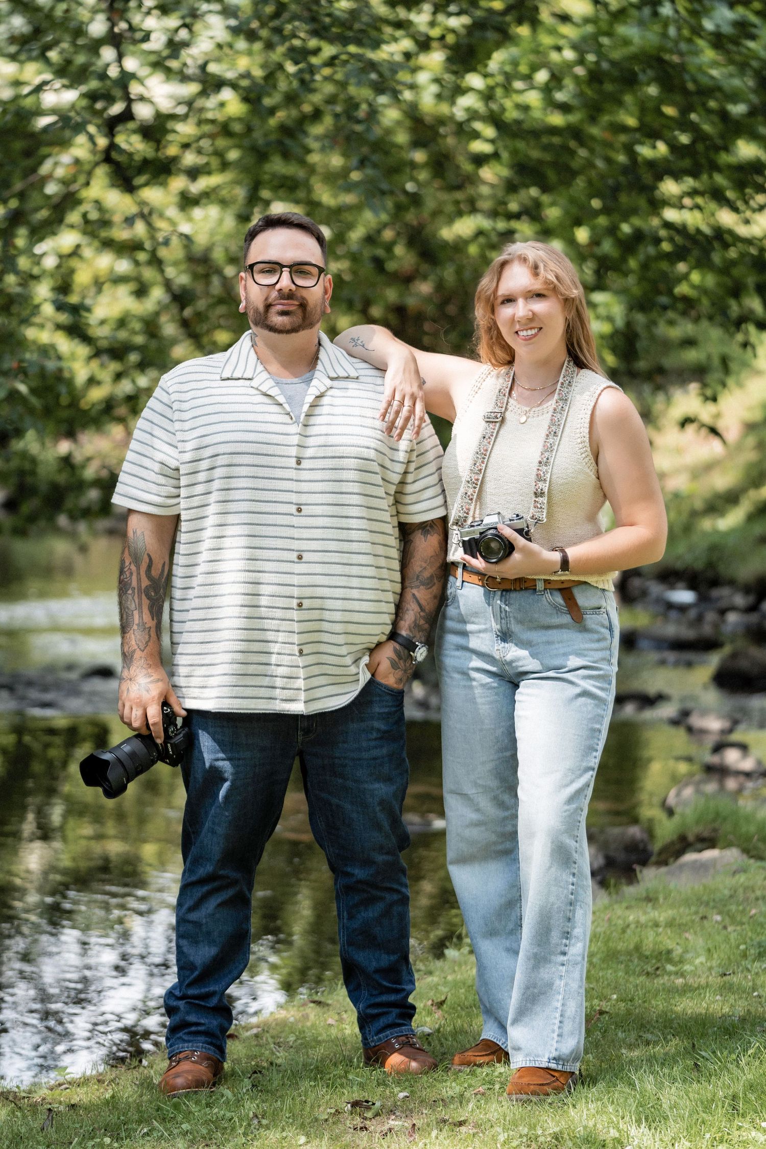 A couple stands together on grass near a pond holding a vintage camera on a sunny summer day.