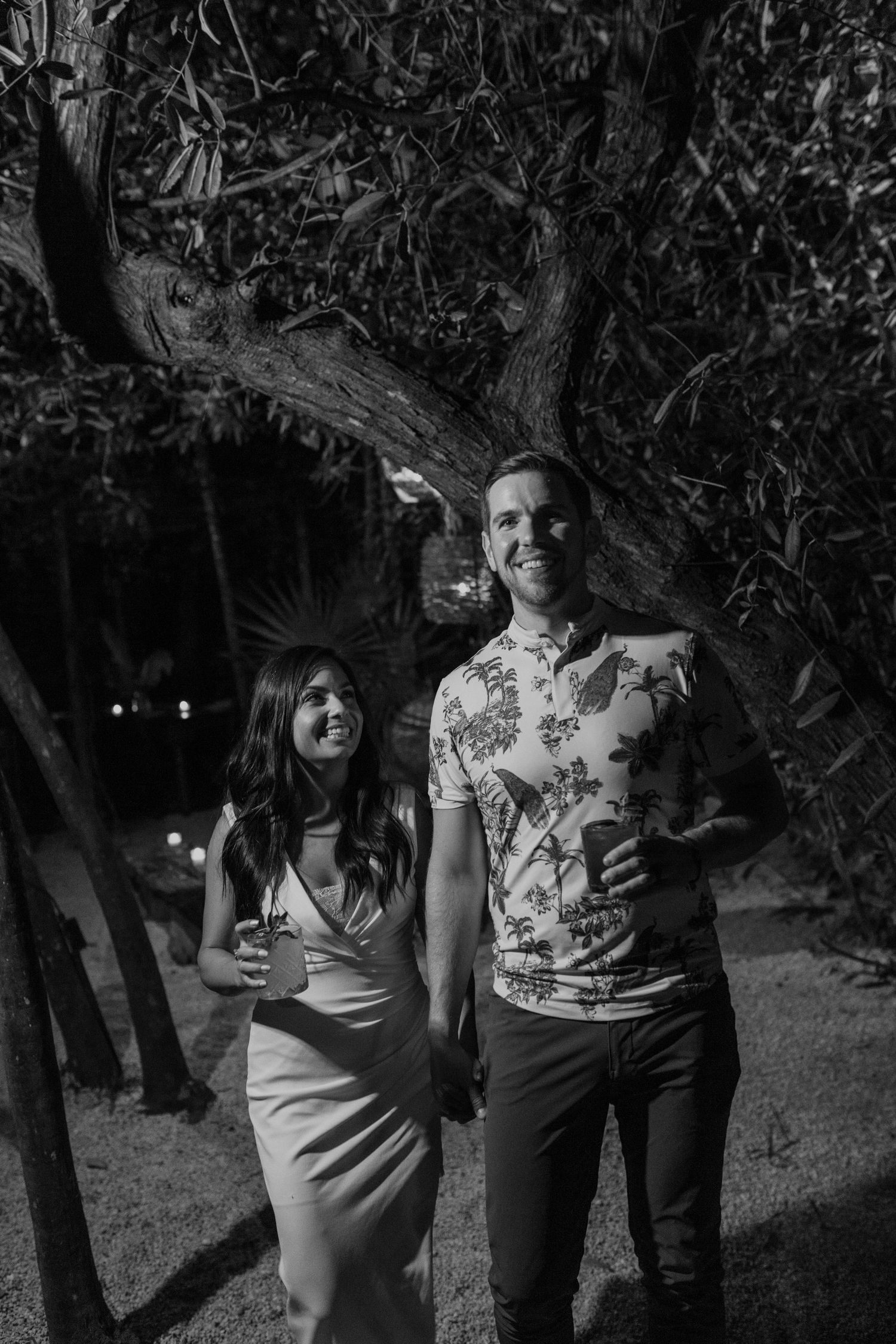 A couple poses together at night under a tree in a candid black and white photo.