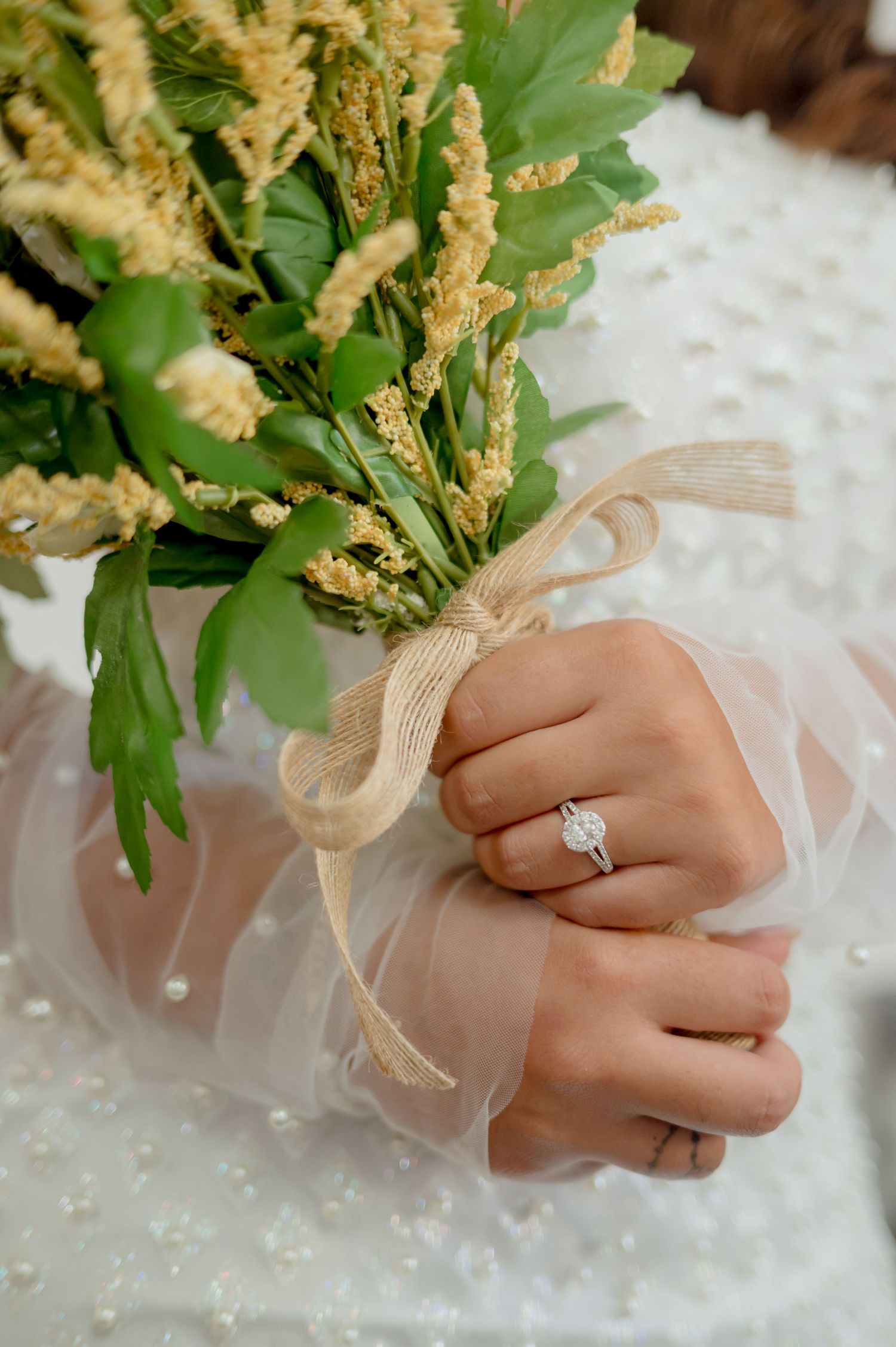 A hand wearing a delicate sheer sleeve and diamond ring holds a bouquet of cream-colored floral stems.