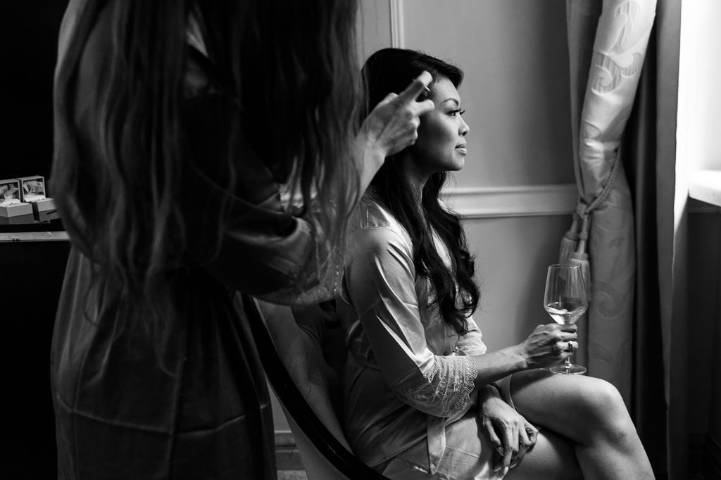 Person drinking from a wine glass while standing near a window in a dark room with natural light coming in.