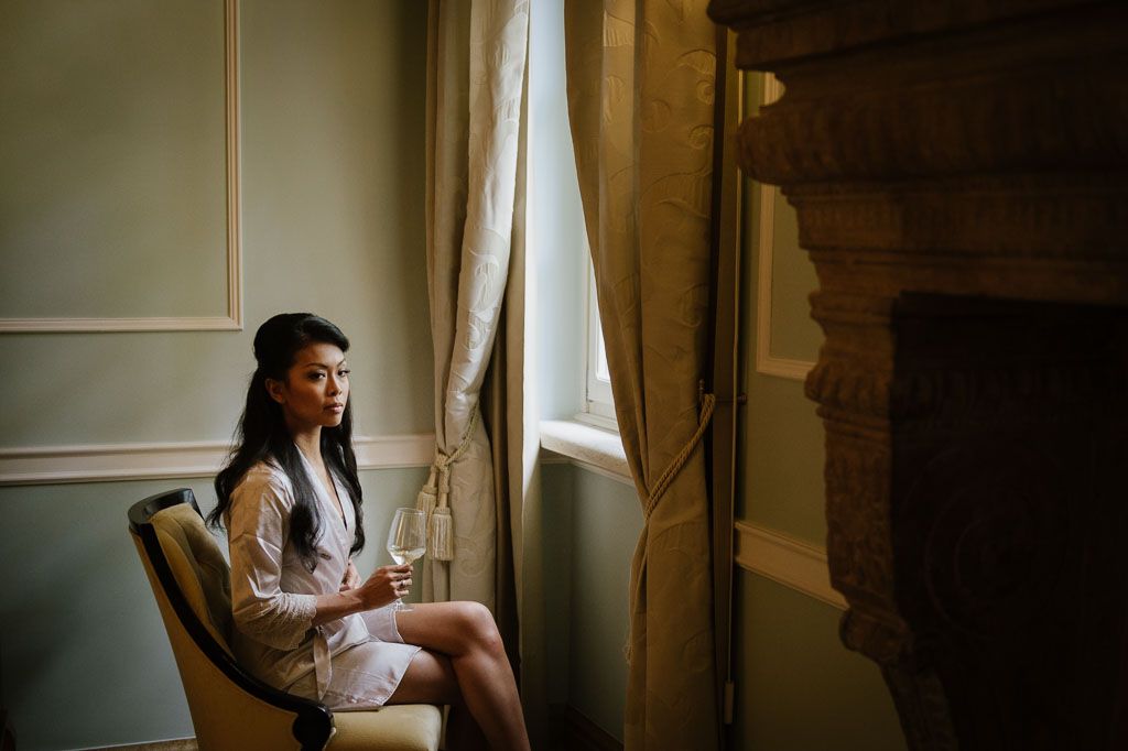 A person sits thoughtfully by a window in a classic interior, holding a beverage while natural light streams in.