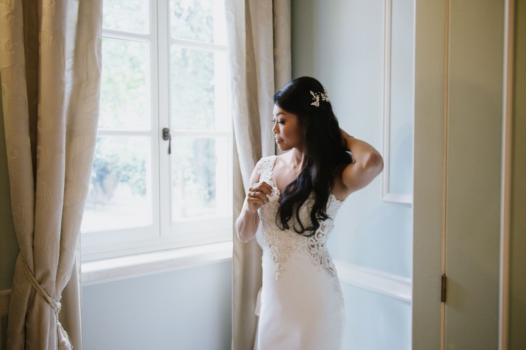 Sunlight streams through windows as a bride adjusts her jewelry in a bright, elegant room.