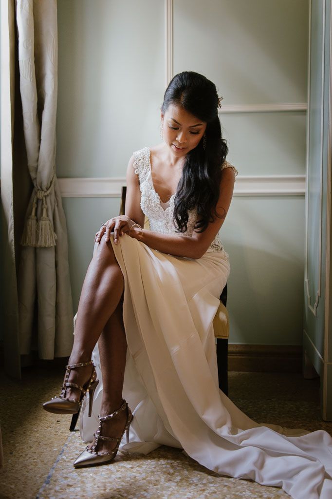 A person in a white wedding dress adjusts their brown heeled shoes while sitting in an elegant room.