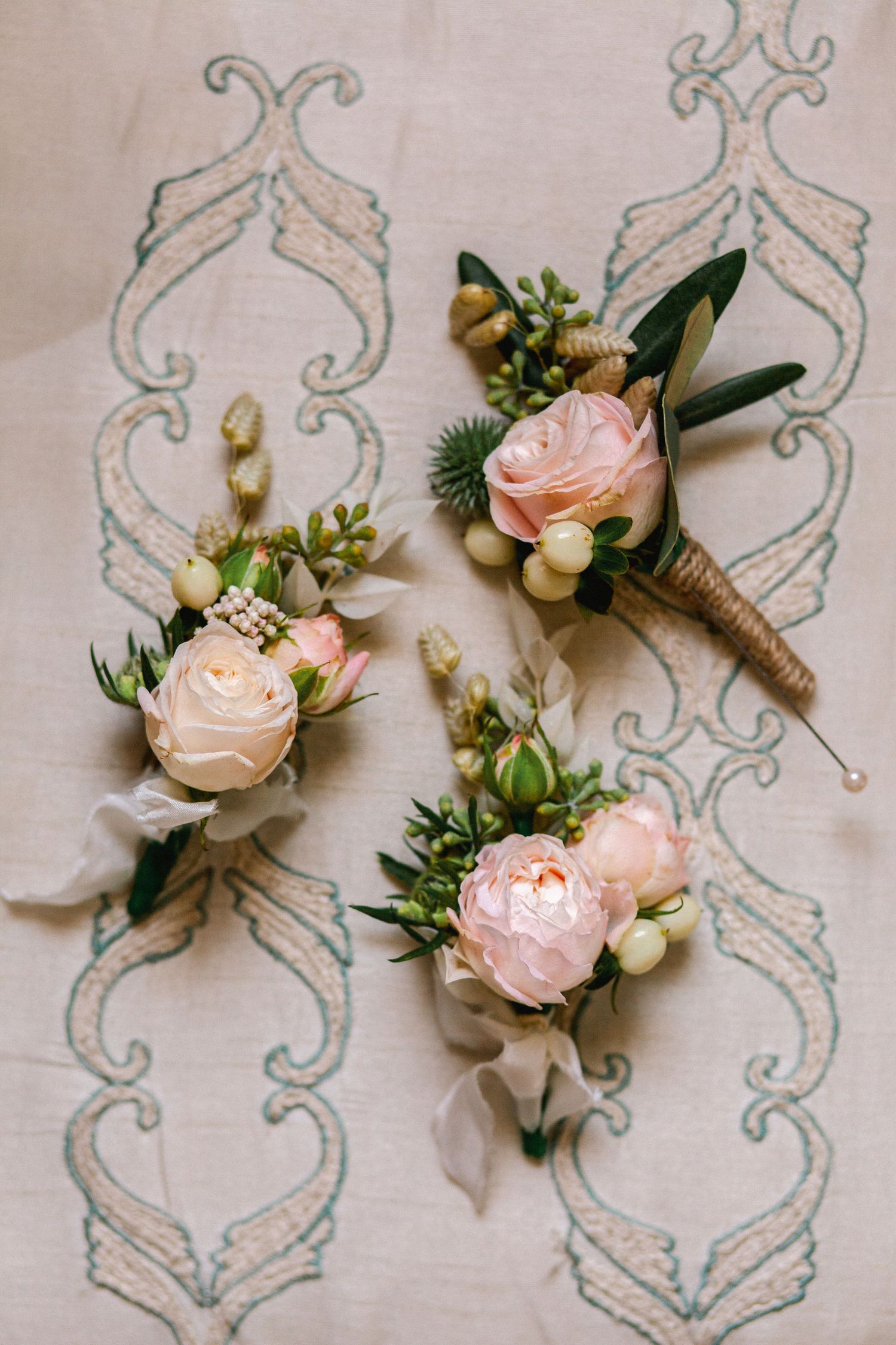 Blush pink rose boutonnieres with greenery arranged on embroidered fabric.