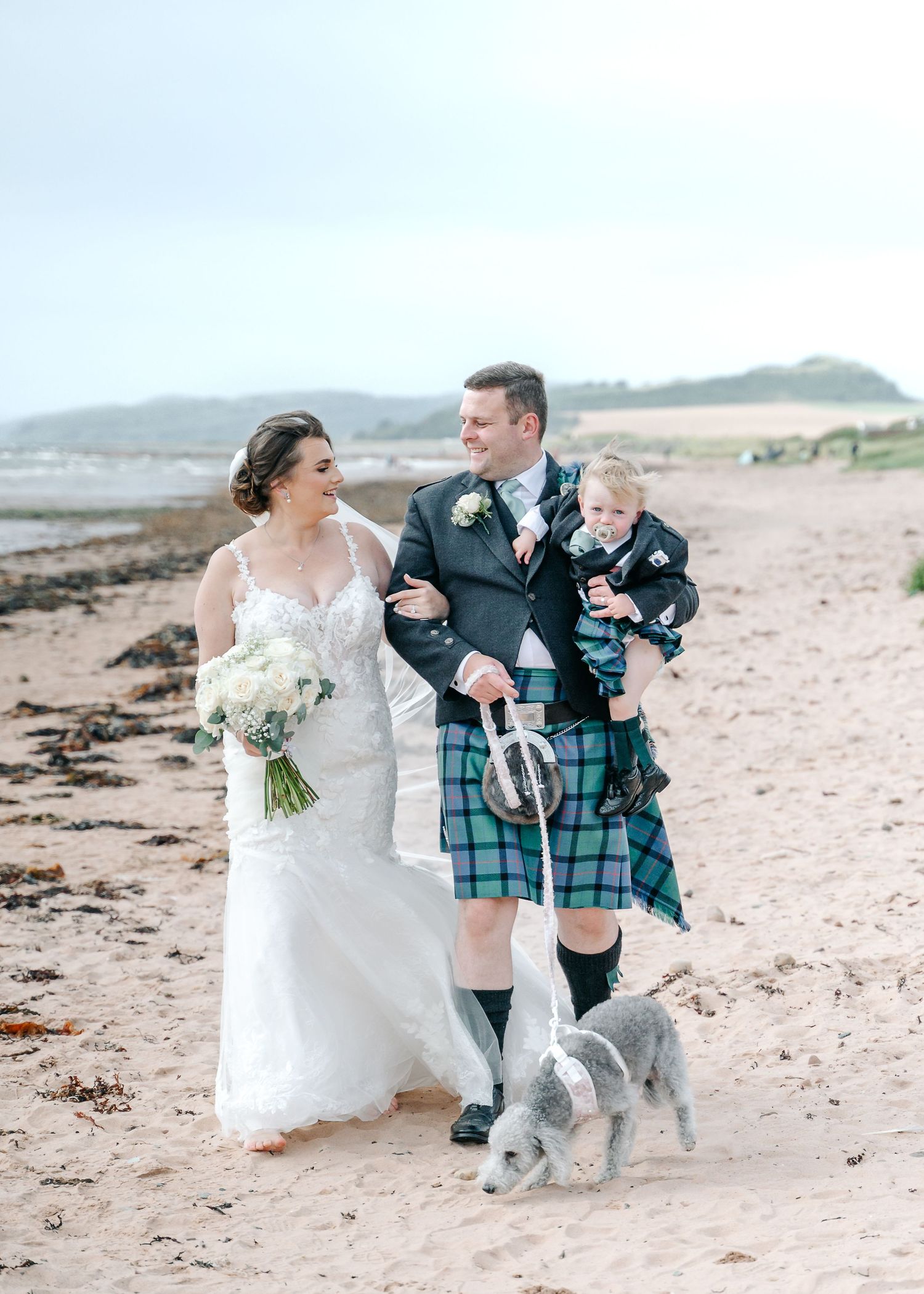 Beach wedding scene with couple in Scottish attire walking their dog along the shoreline.