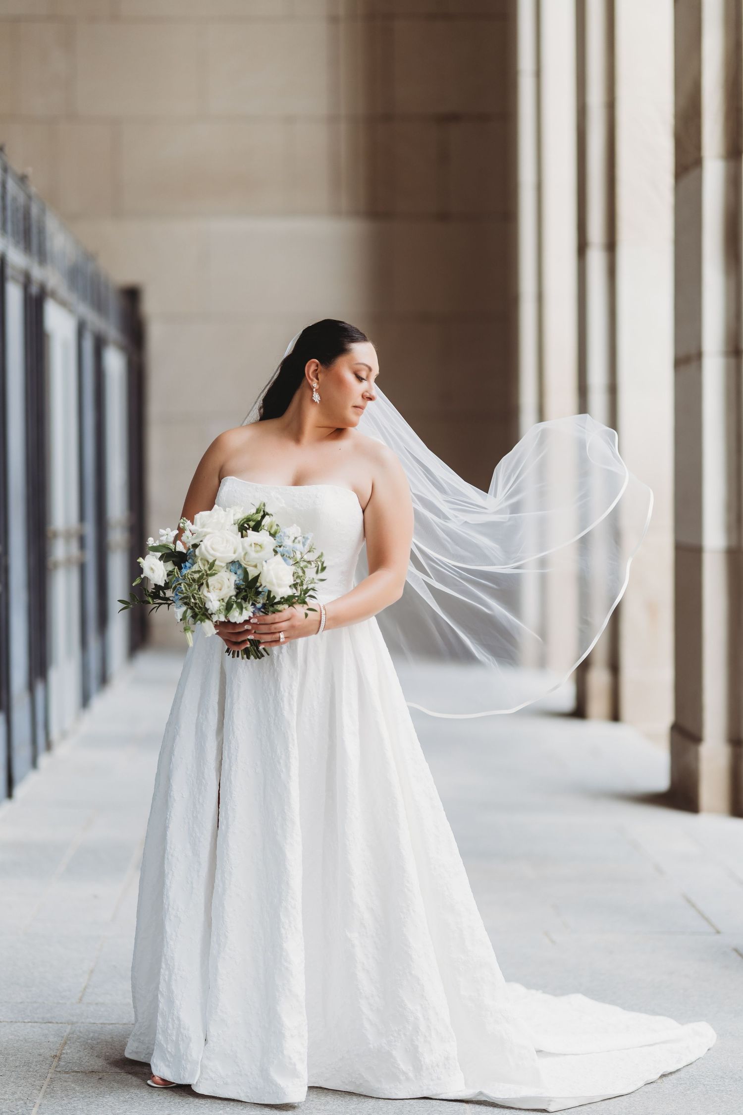 Elegant bride in flowing white gown holds a bouquet while posing in a columned hallway with natural light.