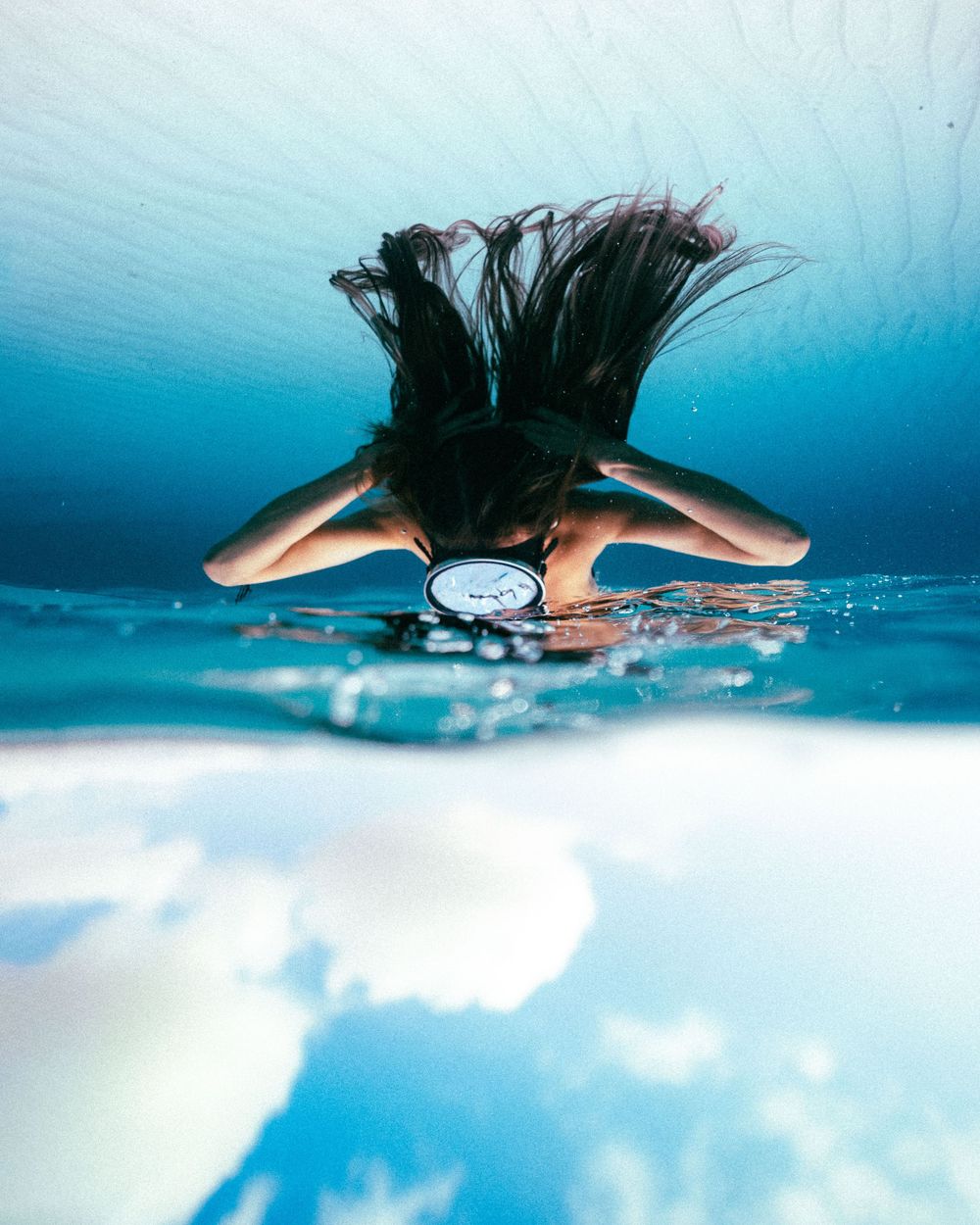 A split-view underwater photo showing a swimmer's dark hair flowing in crystal blue water against a white surface.