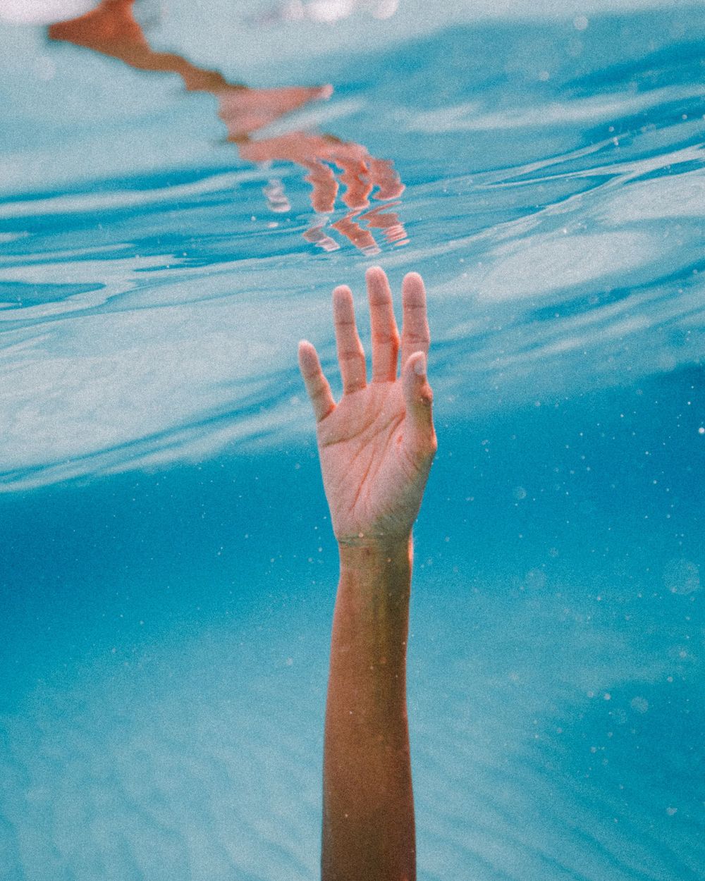 A hand reaches up through turquoise blue water creating ripples on the surface in an artistic underwater photograph.