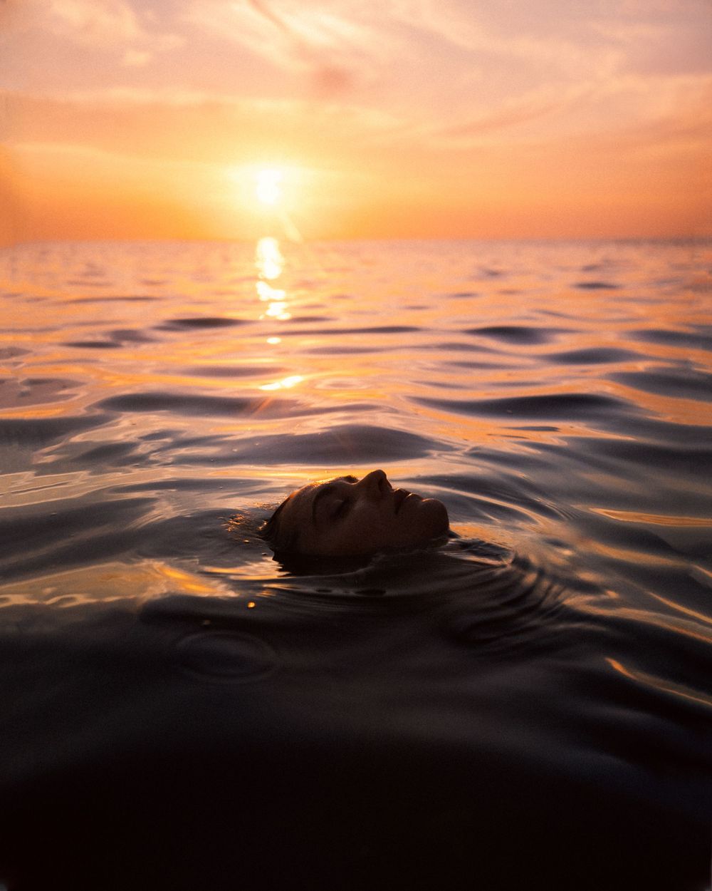 A rock creates gentle ripples in calm ocean waters during a vibrant orange sunset.