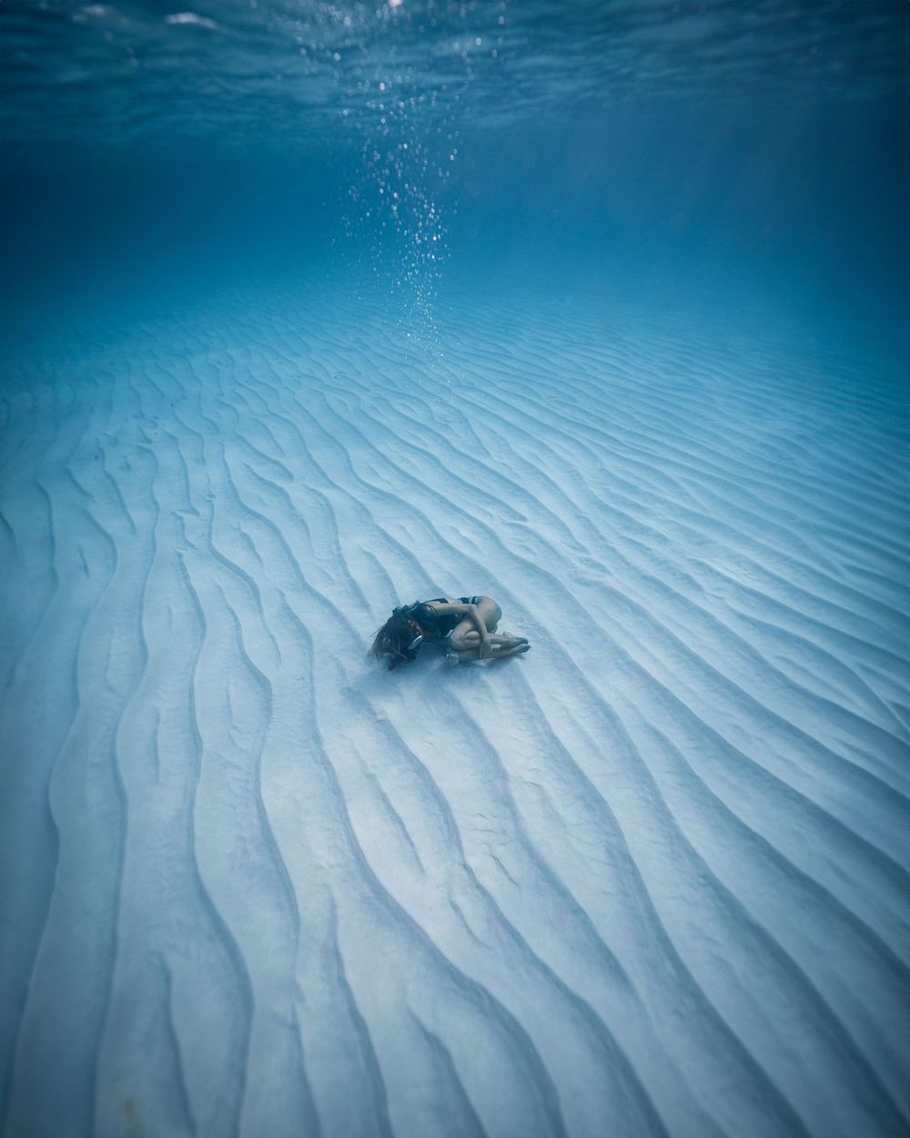 Underwater view of a scuba diver exploring rippled sandy seabed with bubbles rising in clear blue tropical waters.