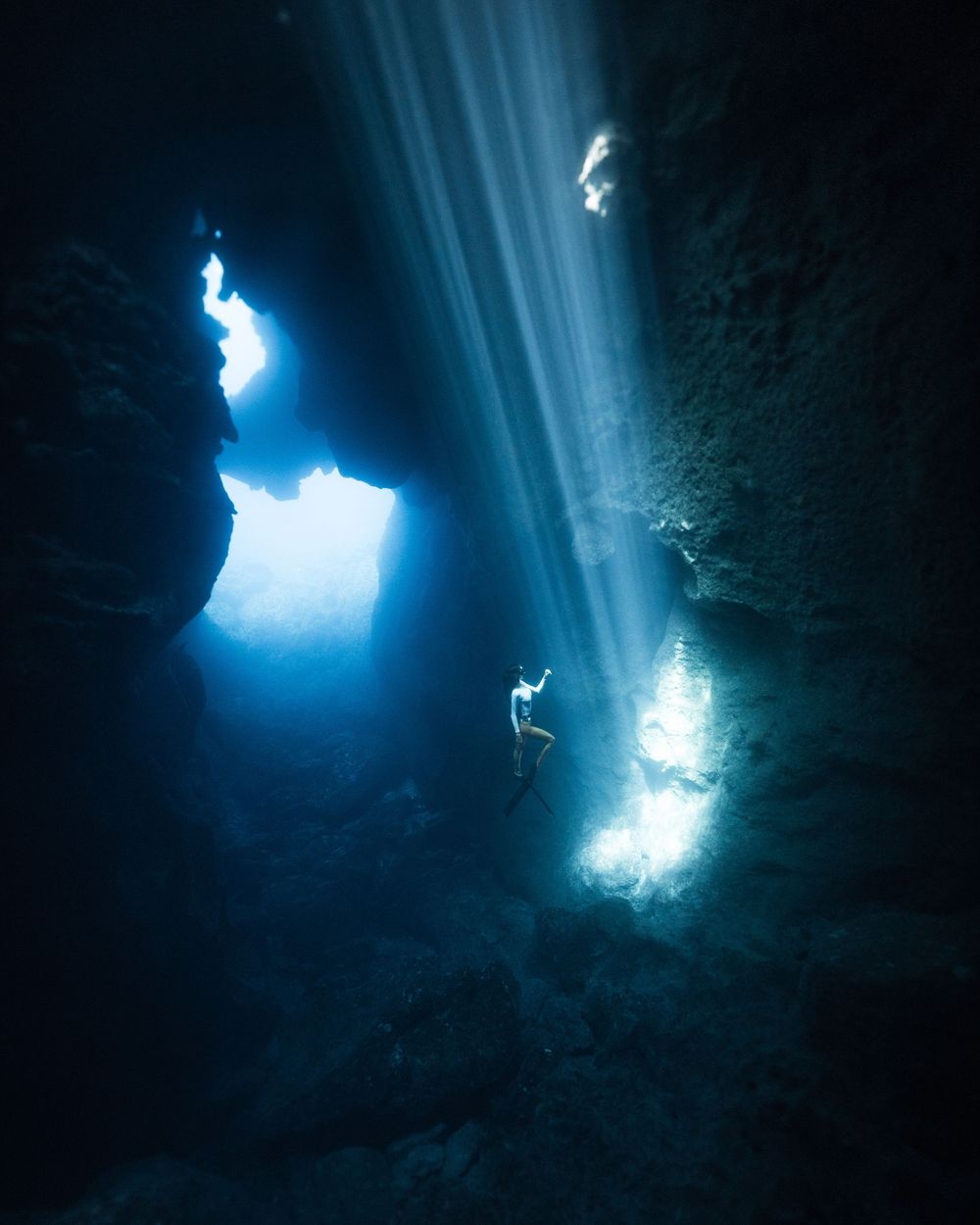Sunlight beams illuminate an underwater cave with crystal clear blue water and rocky formations.