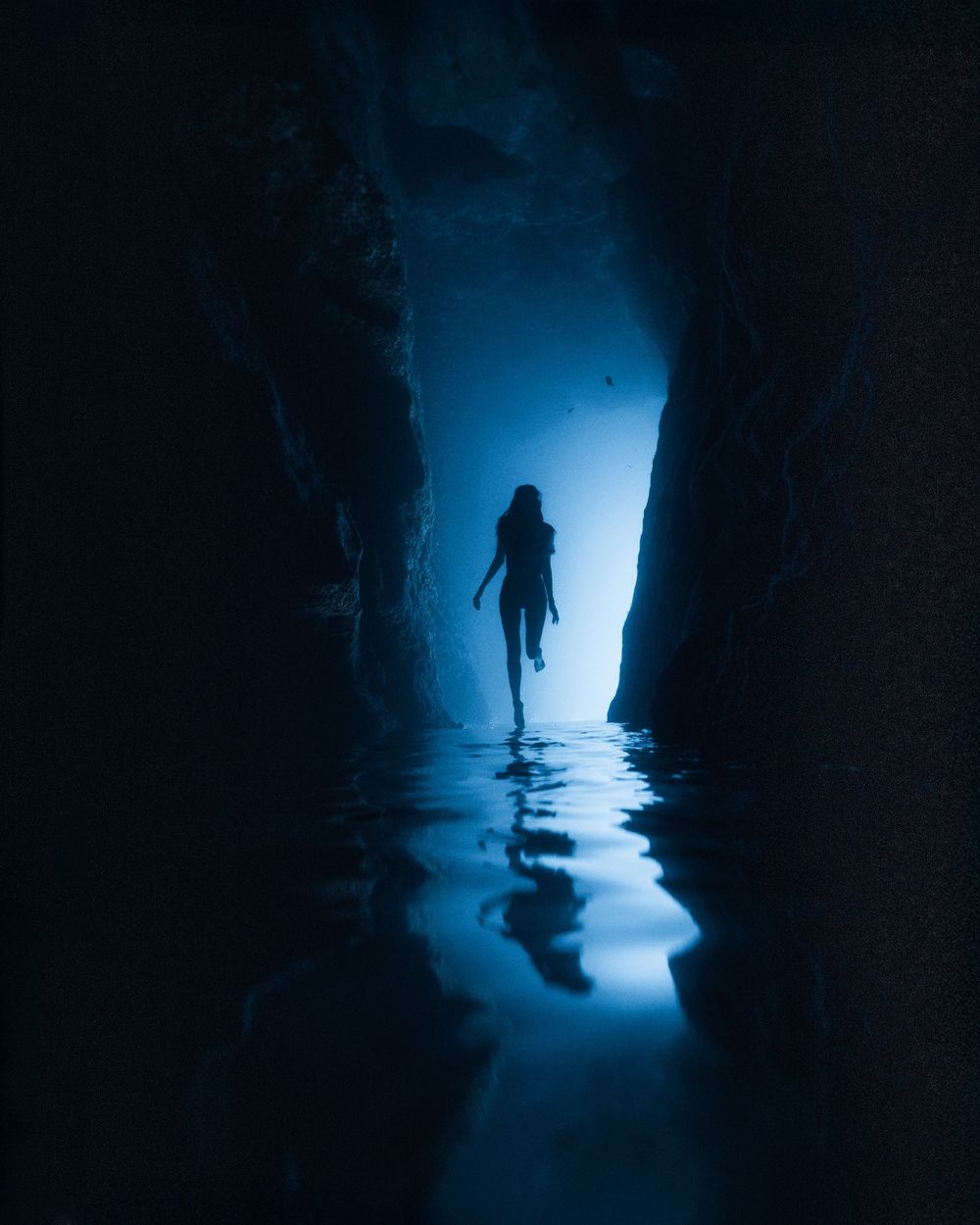 Silhouetted figure walks through dark tunnel with ethereal blue backlighting creating mysterious atmosphere.