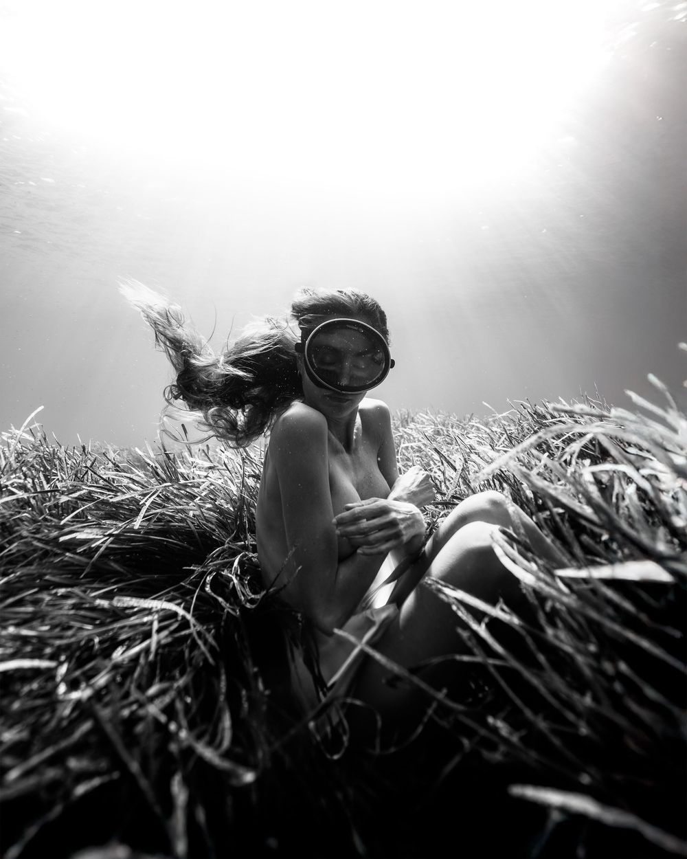 Black and white underwater photo of a woman in a diving mask sitting among sea grass with light rays above.