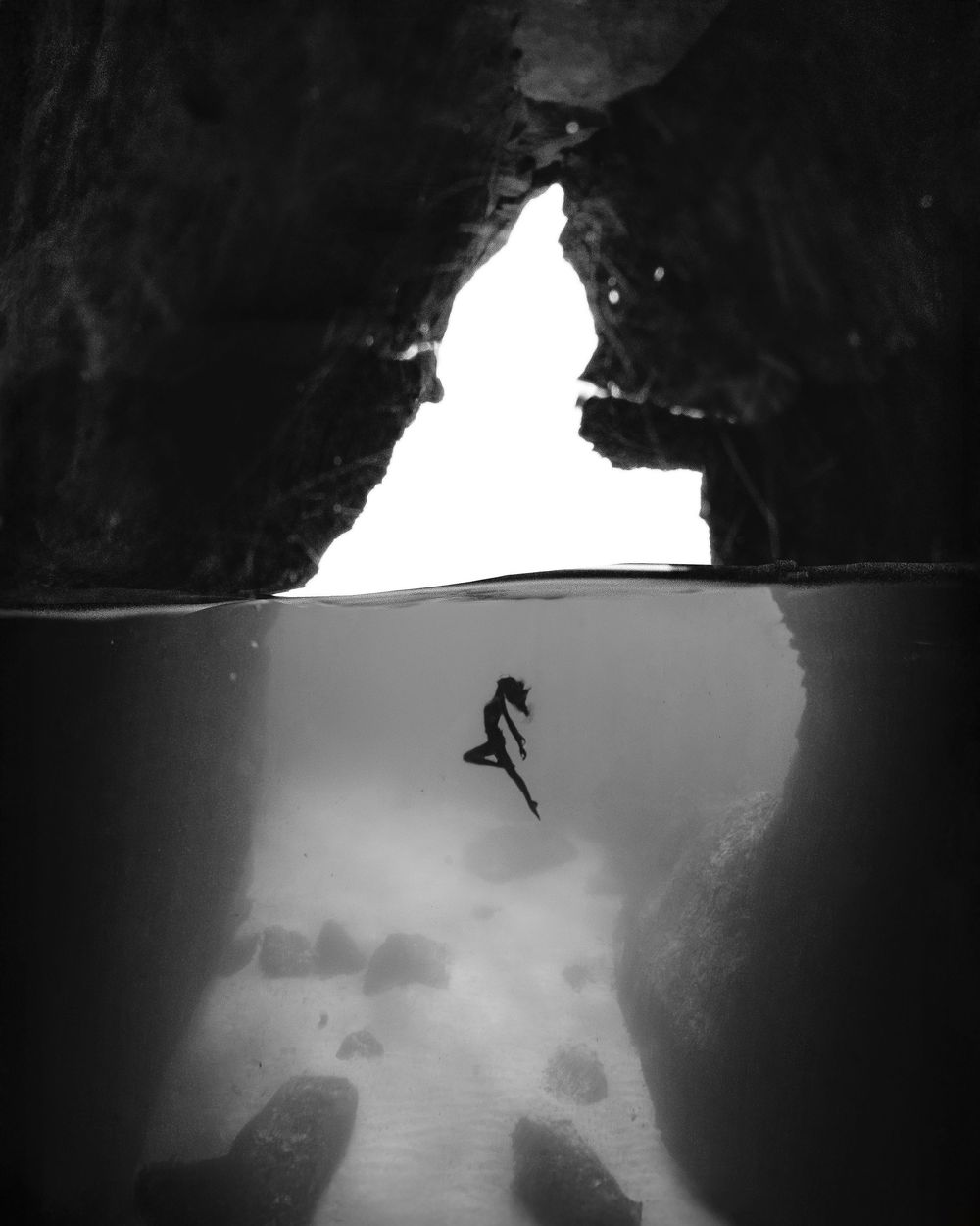 Silhouetted figure diving into dark underwater cave creates dramatic contrast against bright opening in black and white photo.