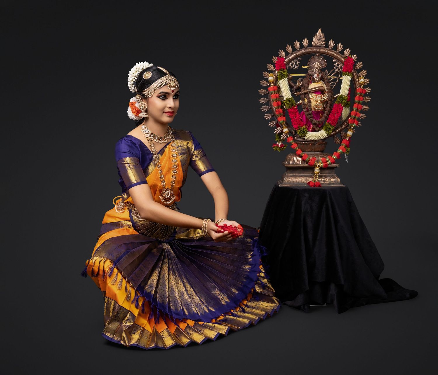 Classical Indian dancer in traditional purple and gold bharatanatyam costume poses beside ornate Nataraja statue.