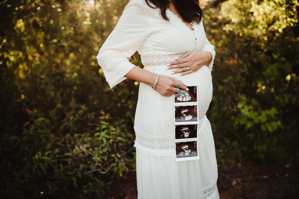 People in white clothing holding ultrasound photos during a sunset maternity photoshoot in a natural outdoor setting.