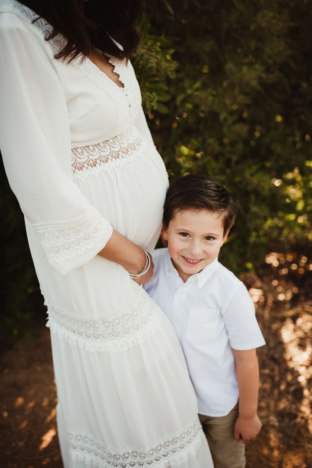 A young child in white stands next to someone wearing a white lace dress in a natural outdoor setting.