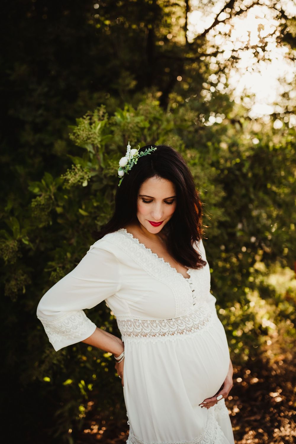 At sunset a person poses in a flowing white dress with lace details against a backdrop of trees and golden light.