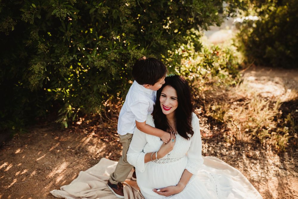 A couple in white clothing embraces while sitting on the ground in warm sunlit nature setting.